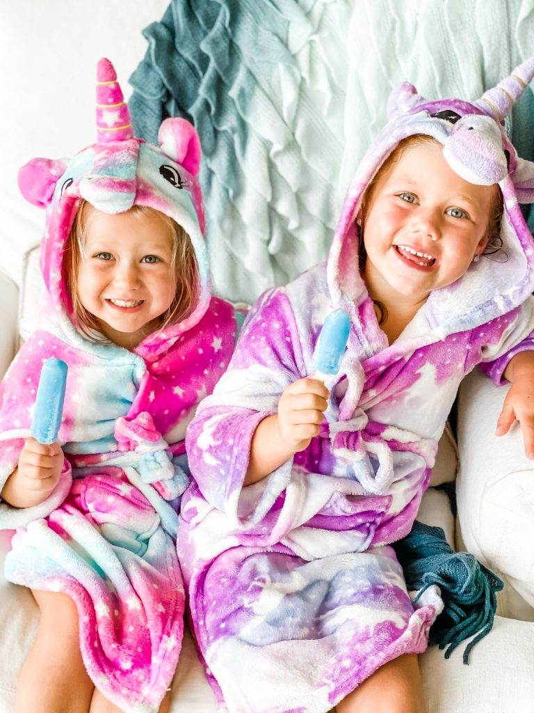 Two children wrapped in unicorn robes, holding blue popsicles. 